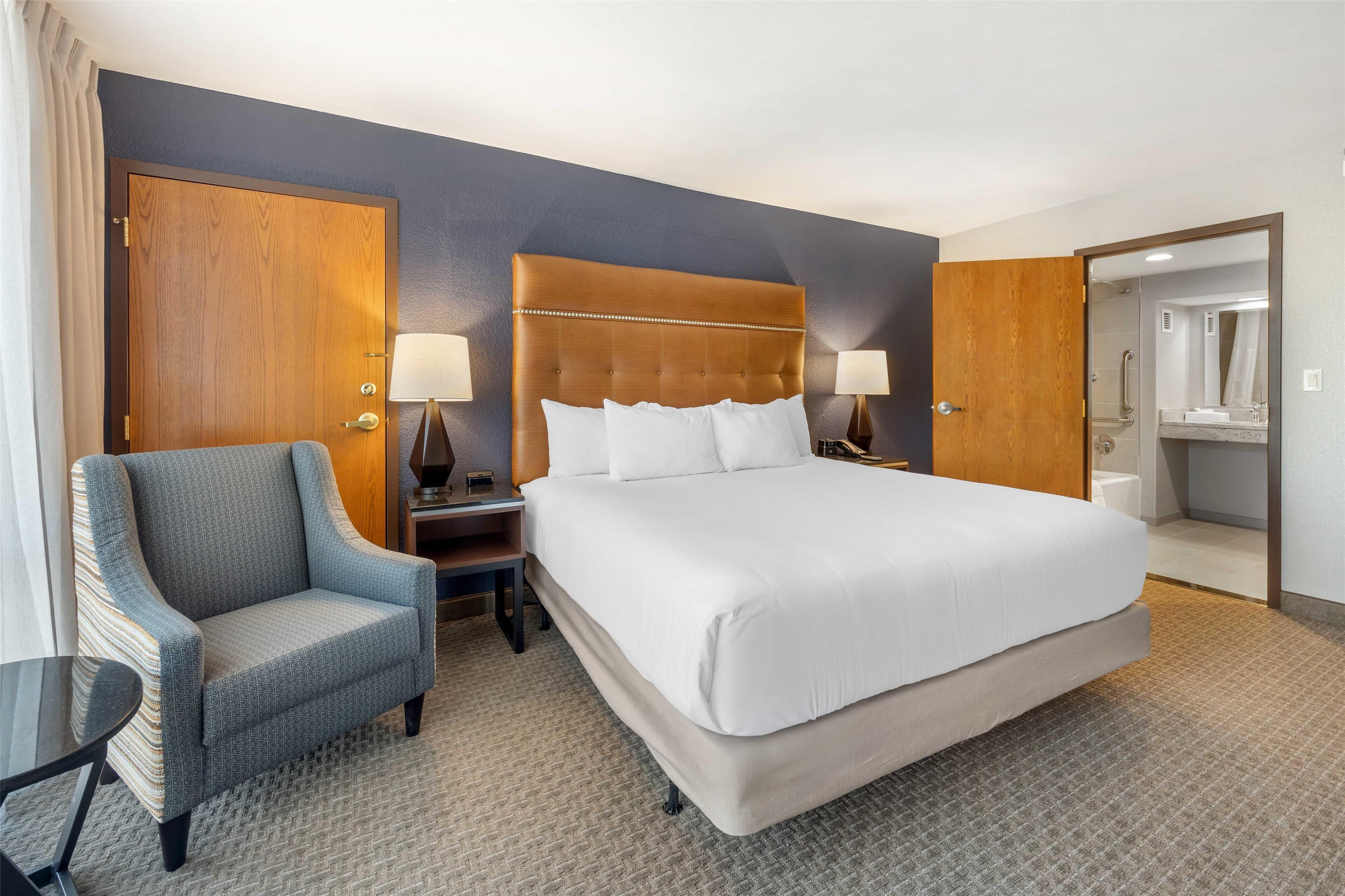 Ein Hotelzimmer mit einem großen Bett, einem Sessel, einem Nachttisch, einer Lampe und einem durchgegangene Tür erkennbaren Badezimmer.