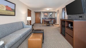 A hotel room with a sofa, a small table, a bench, a dining area with chairs, a kitchen area with a microwave, and a flat-screen TV mounted on the wall.