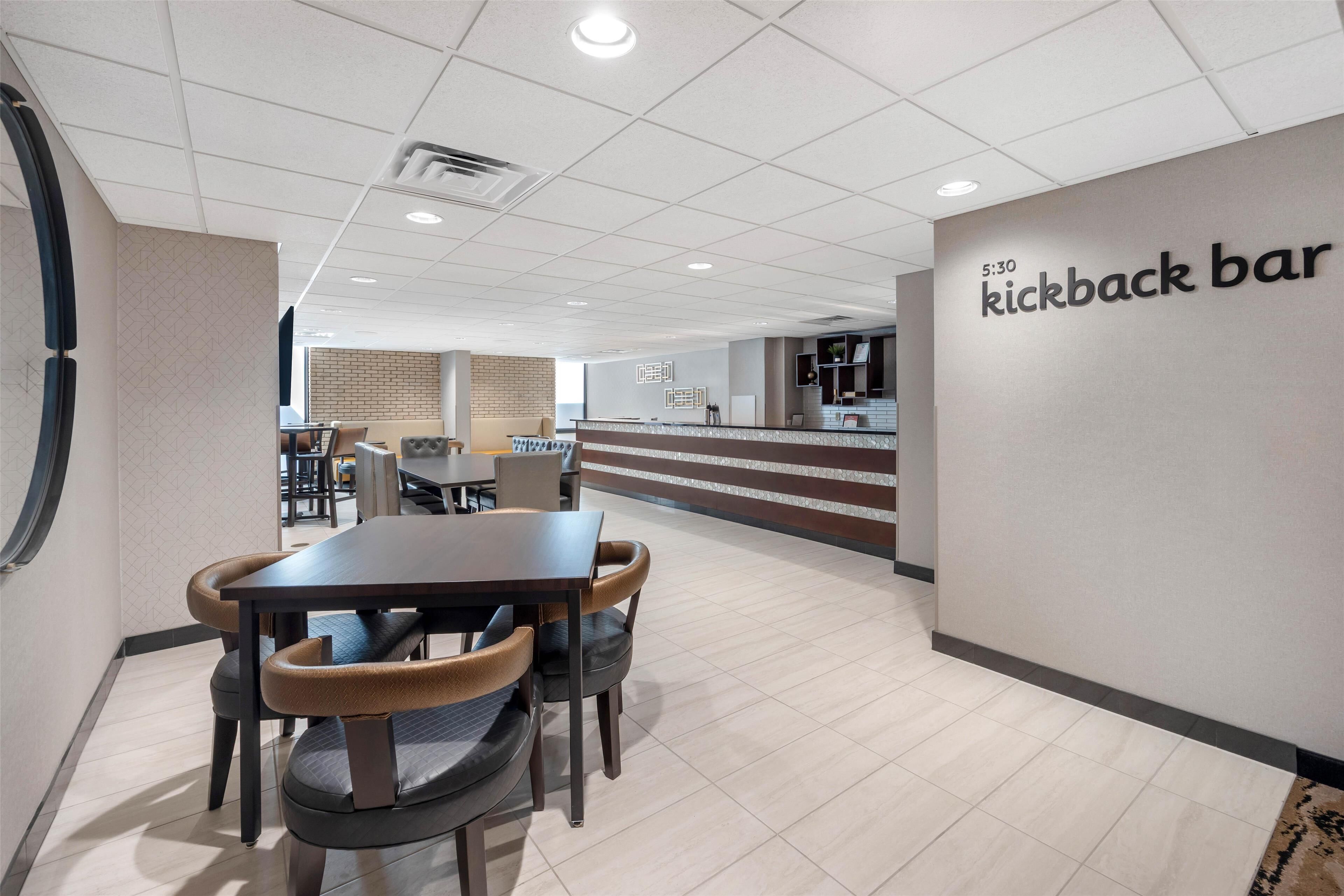 Un interior moderno con una zona de comedor que incluye una mesa y sillas, un mostrador de recepción y una pared con un cartel que dice "kickback bar".