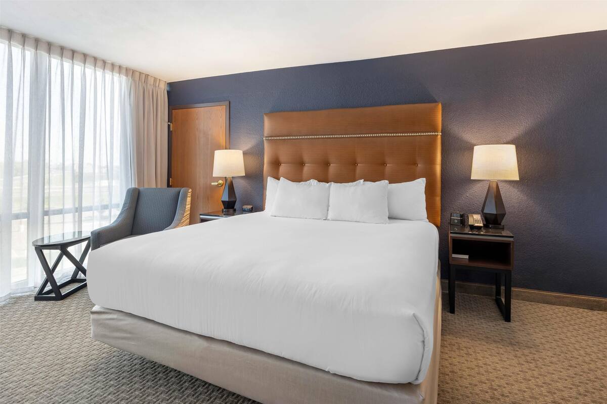 Una cama grande con ropa blanca, dos mesitas de noche con lámparas, una silla y vistas al exterior a través de cortinas translúcidas.