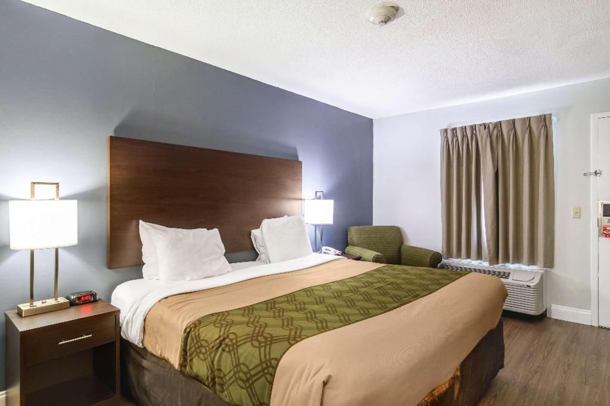 A hotel room with a bed, bedside table, lamp, chair, and a window with curtains.