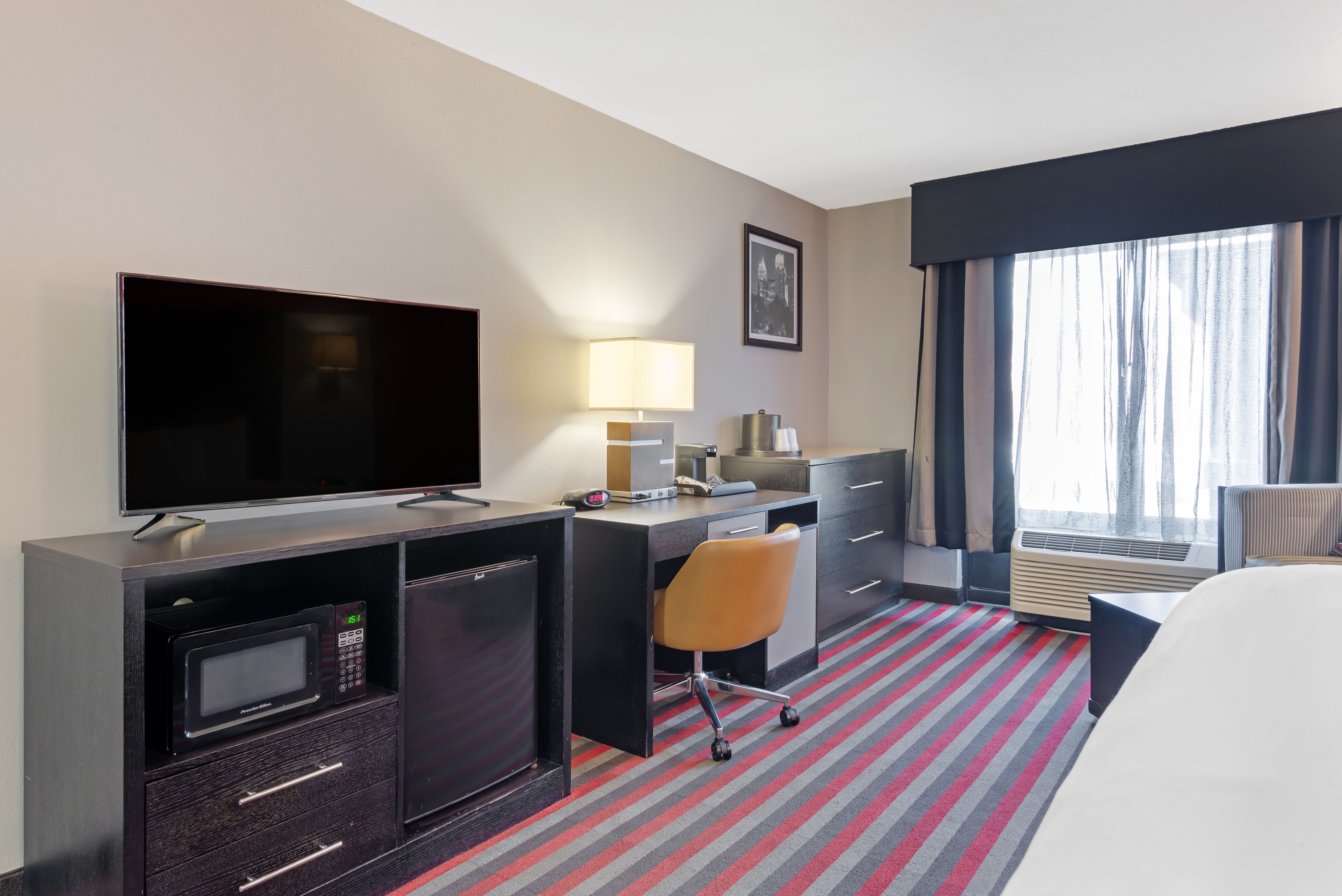 A hotel room with a flat-screen TV, a desk with a lamp, a microwave, a chair, and a large window with curtains.