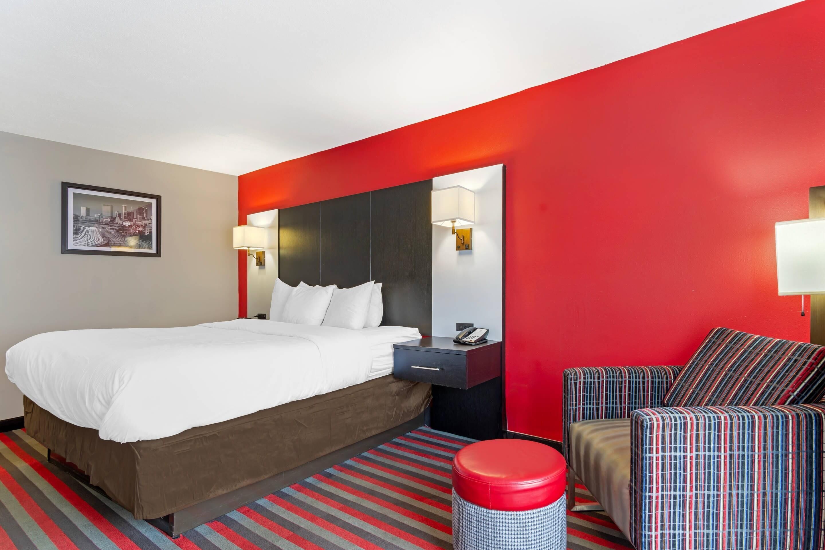 A hotel room with a large bed, a red accent wall, a striped rug, a striped chair, and a nightstand with a lamp.