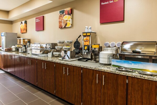 A breakfast bar with various food and drink stations, including a waffle maker, coffee machines, and a juice dispenser.