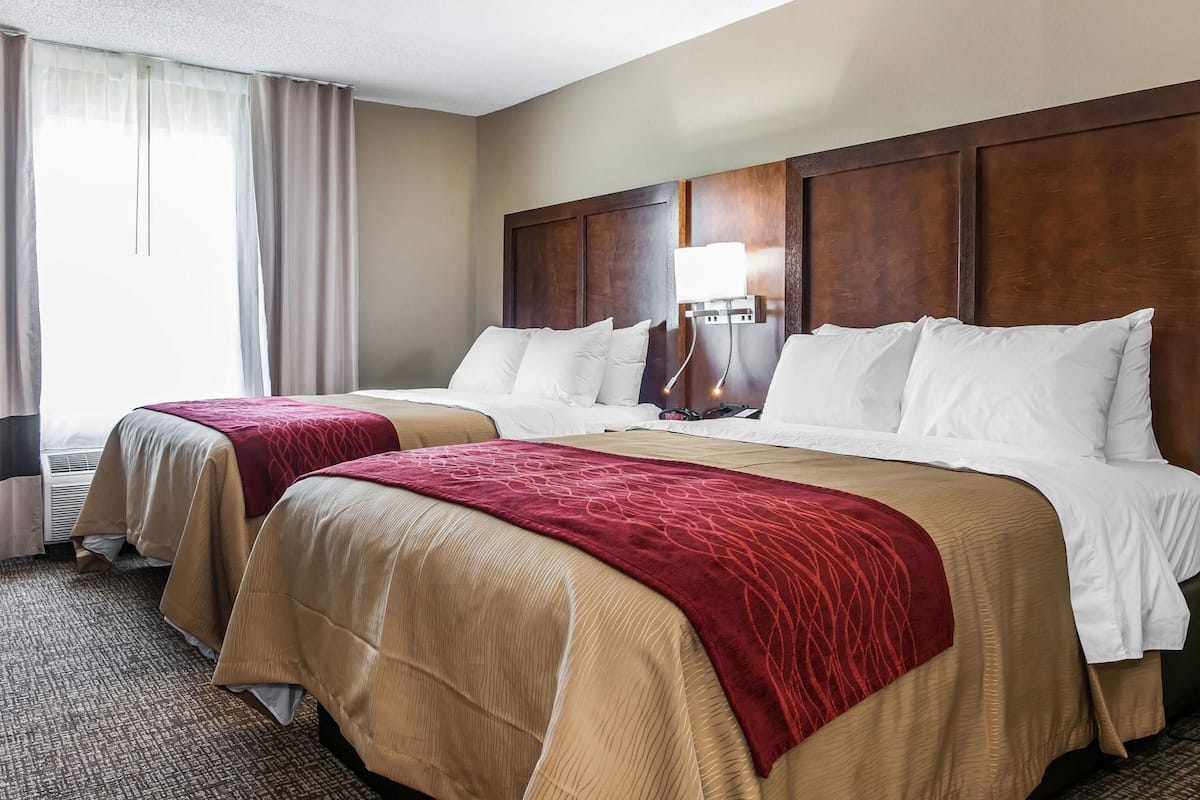 A hotel room with two beds, beige and red bedspreads, a wooden headboard, a bedside table with a lamp, and a window with curtains.