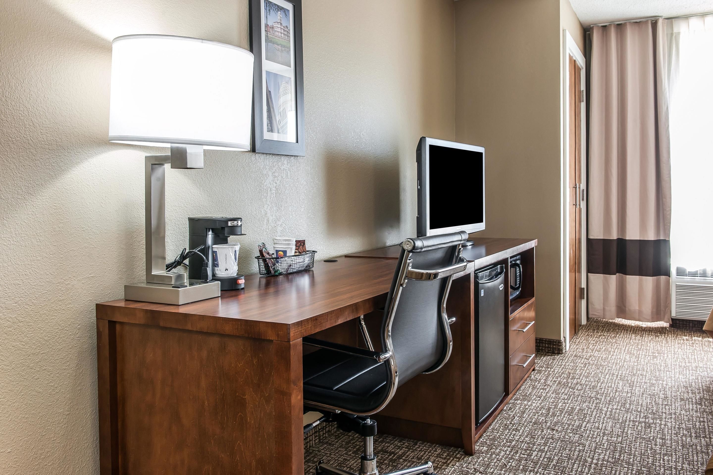 Habitación de hotel con un escritorio de madera, una silla de oficina negra, un televisor de pantalla plana, una cafetera y un cuadro en la pared.