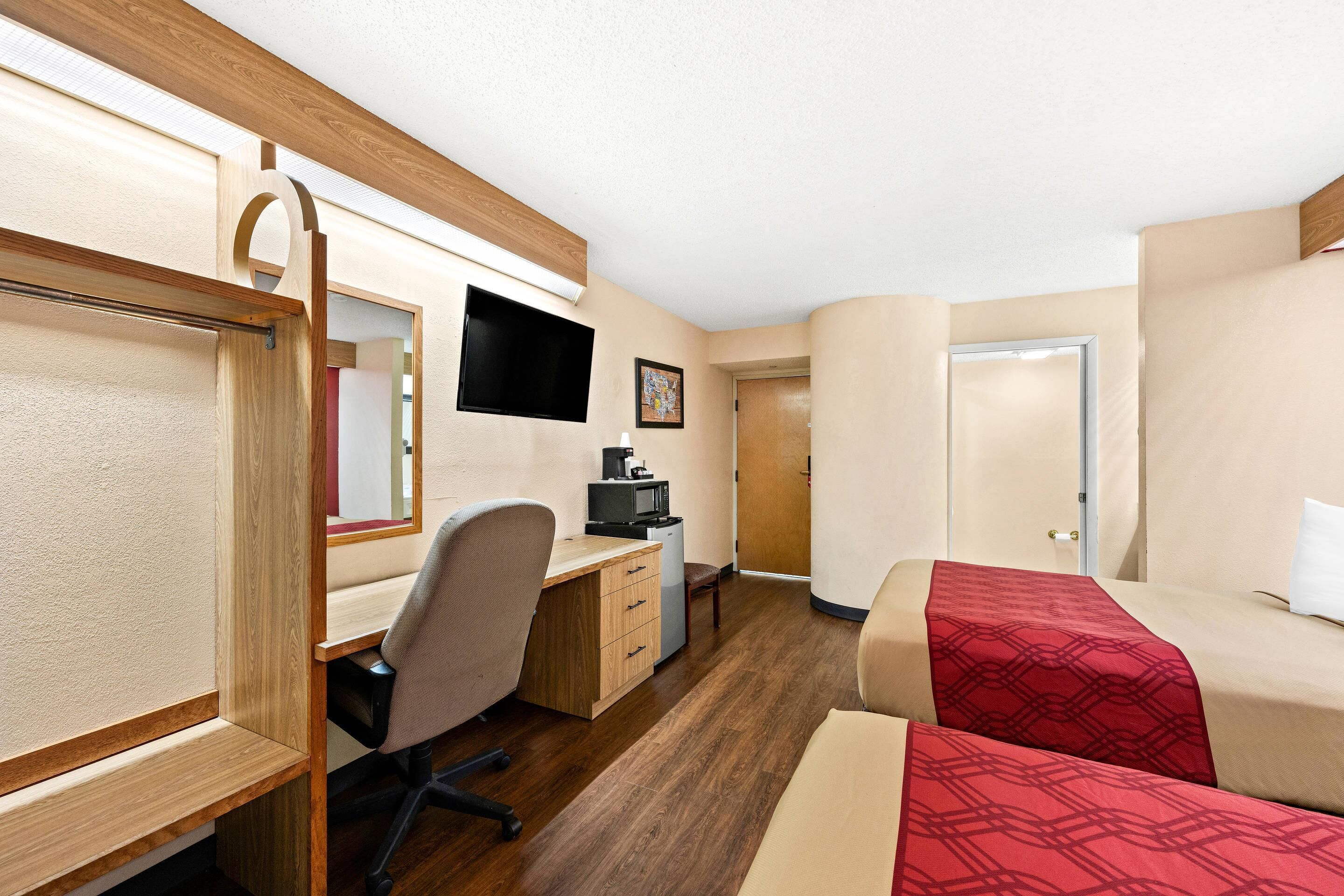 A hotel room with a wooden desk, a chair, a bed with a red blanket, a television, and a door.