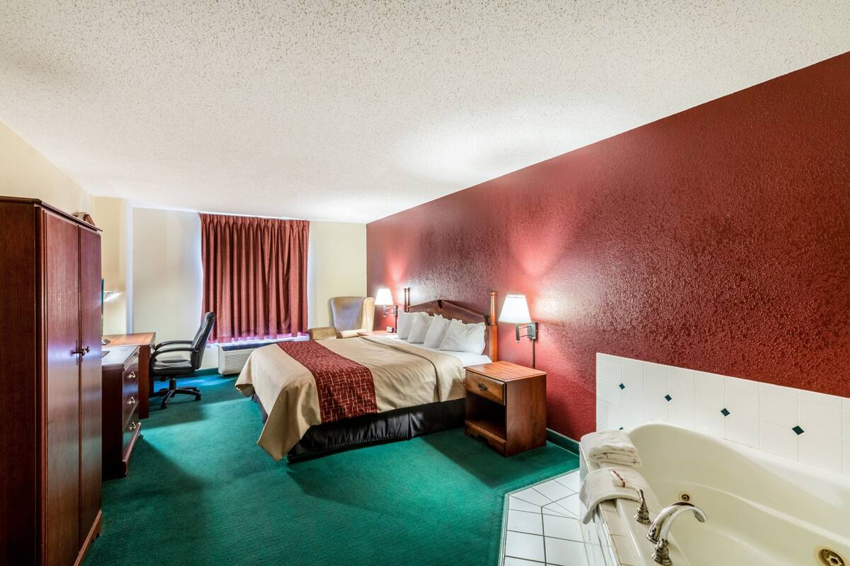 A hotel room with a bed, a desk, a chair, a nightstand, a lamp, and a bathtub.