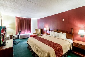 Una habitación de hotel con una cama grande, un escritorio con una computadora, una silla, una ventana con cortinas y una lámpara fijada a la pared.