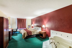 Habitación de hotel con cama, escritorio, silla, mesita de noche, lámpara y bañera.