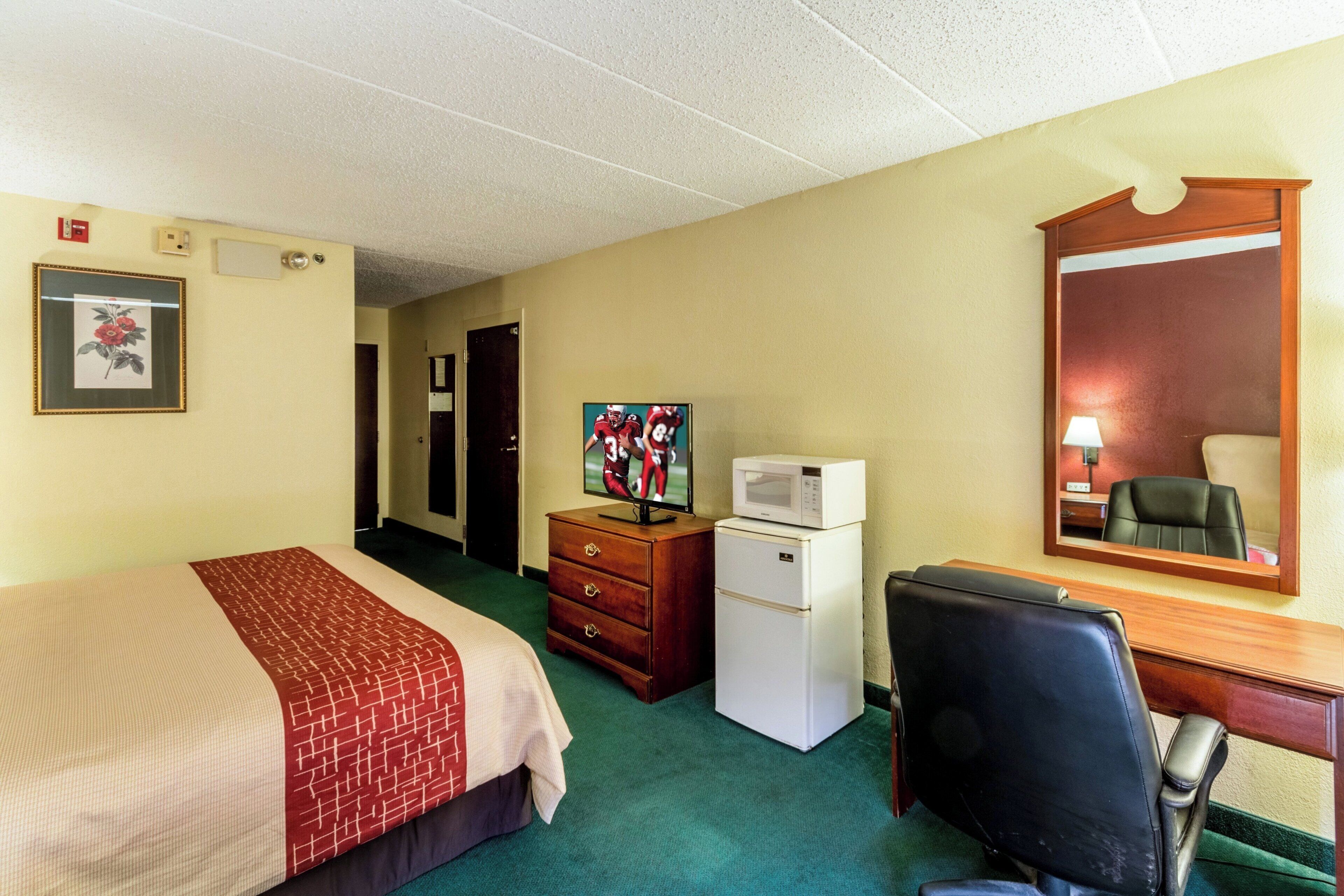 Quarto de hotel com cama, escrivaninha, cadeira, geladeira, televisão e espelho.
