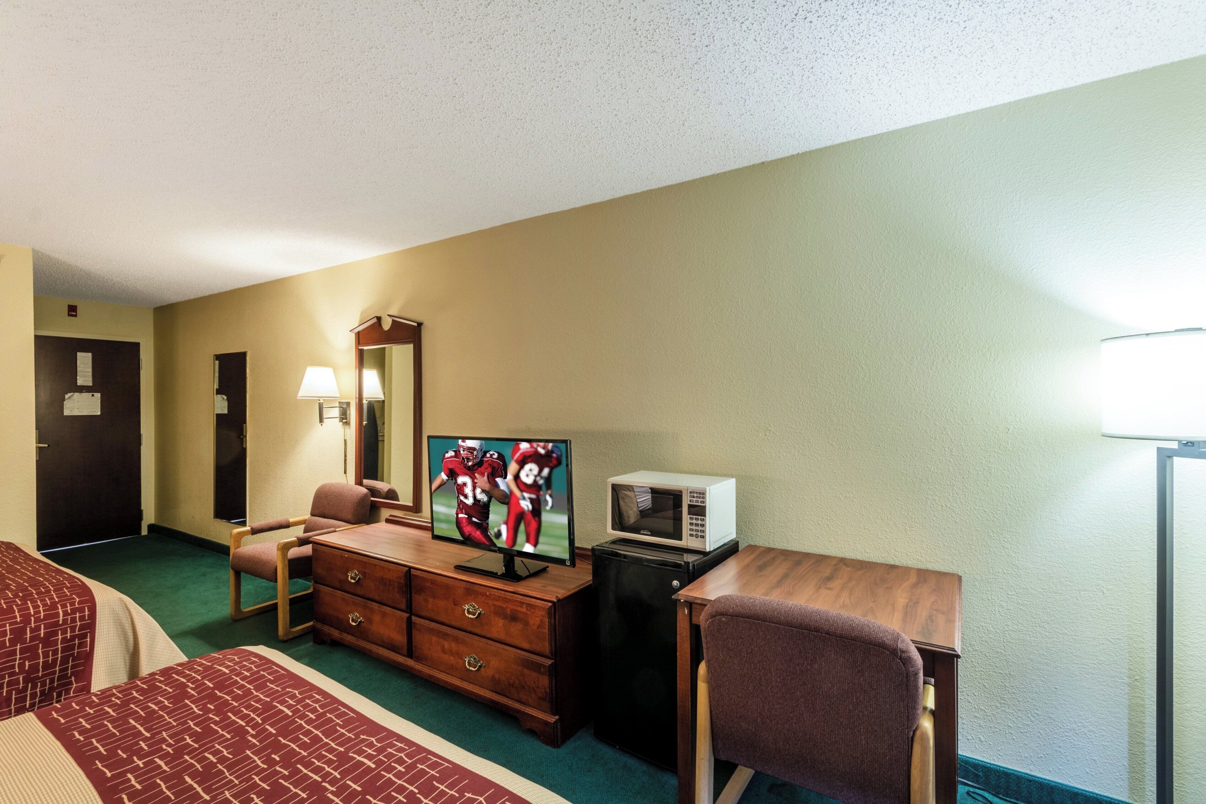 A hotel room with a bed, a desk with a television, a microwave, a dresser, a chair, and a lamp.