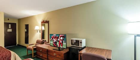 A hotel room with a bed, a desk with a television, a microwave, a dresser, a chair, and a lamp.