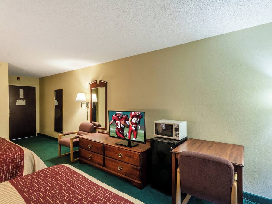 A hotel room with a bed, a desk with a television, a microwave, a dresser, a chair, and a lamp.