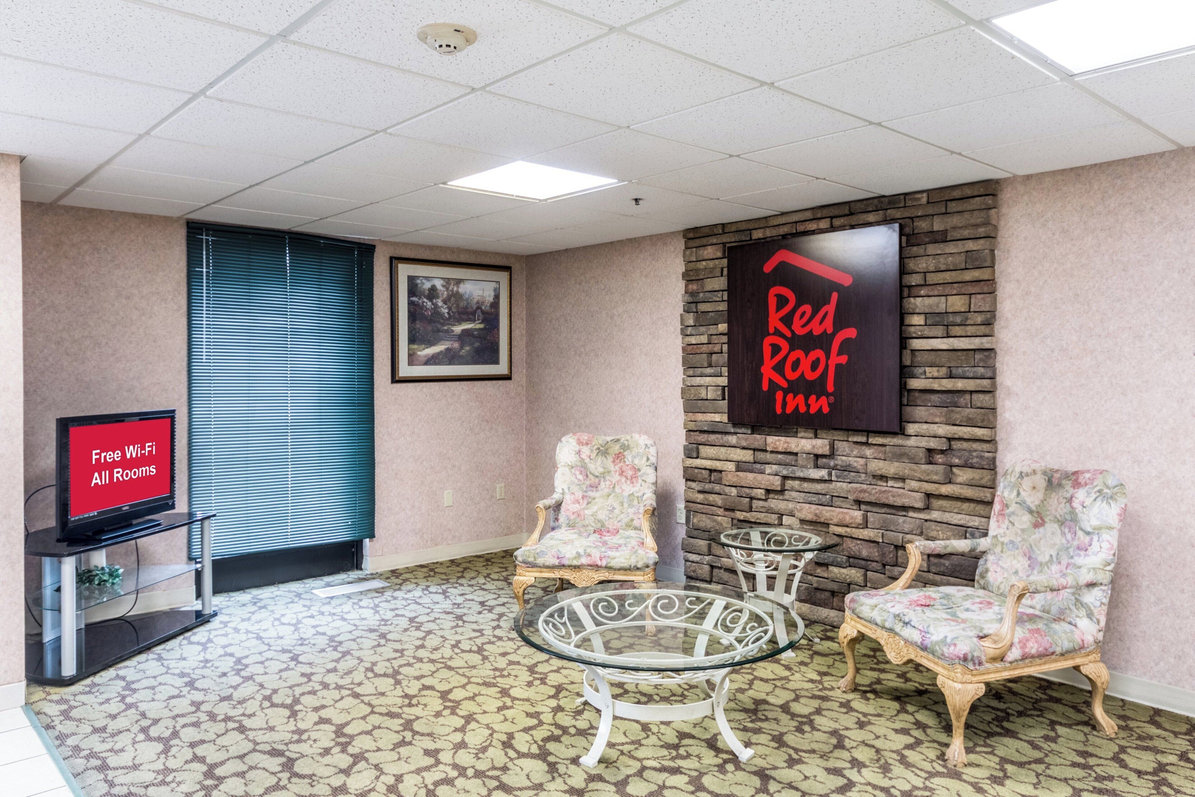 A room with a television displaying 'Free Wi-Fi All Rooms,' a framed painting, a 'Red Roof Inn' sign, and two floral armchairs with a glass table.