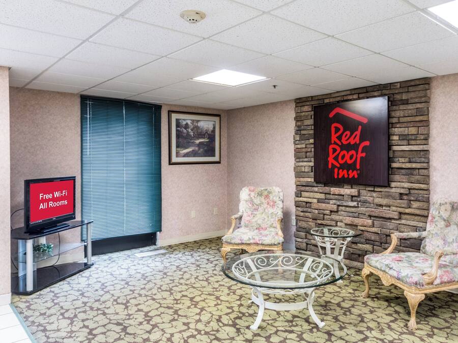 A room with a television displaying 'Free Wi-Fi All Rooms,' a framed painting, a 'Red Roof Inn' sign, and two floral armchairs with a glass table.