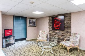 A room with a television displaying 'Free Wi-Fi All Rooms,' a framed painting, a 'Red Roof Inn' sign, and two floral armchairs with a glass table. - Red Roof Inn Morehead (Morehead)