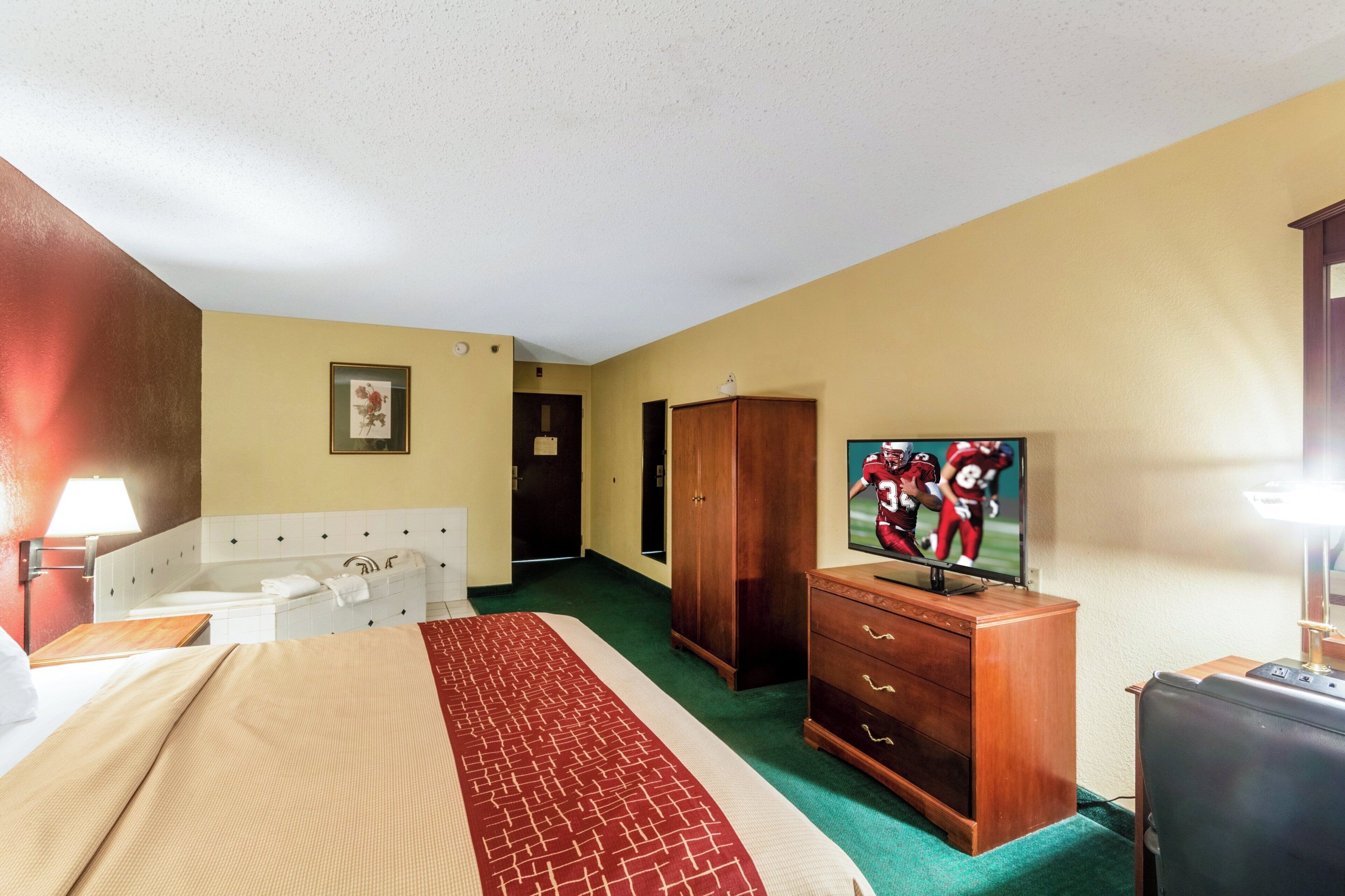 A hotel room with a bed, a television, a wooden dresser, a desk, and a bathroom with a bathtub.