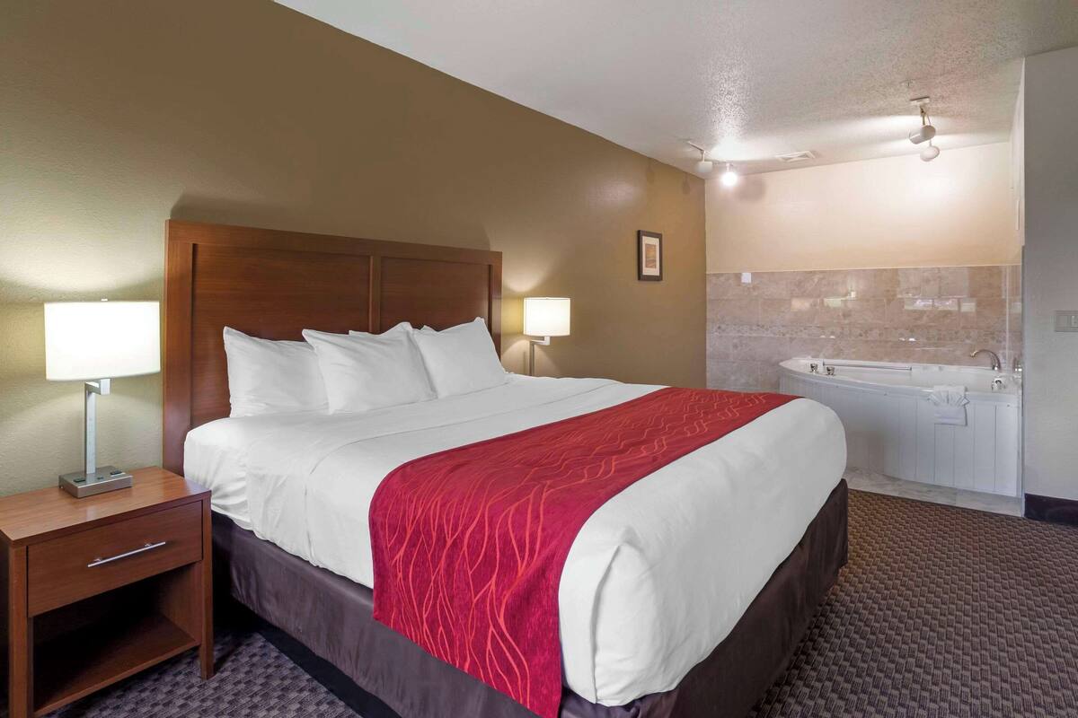 A hotel room with a large bed, a nightstand, a lamp, a red blanket, a bathroom with a bathtub, and a shower.