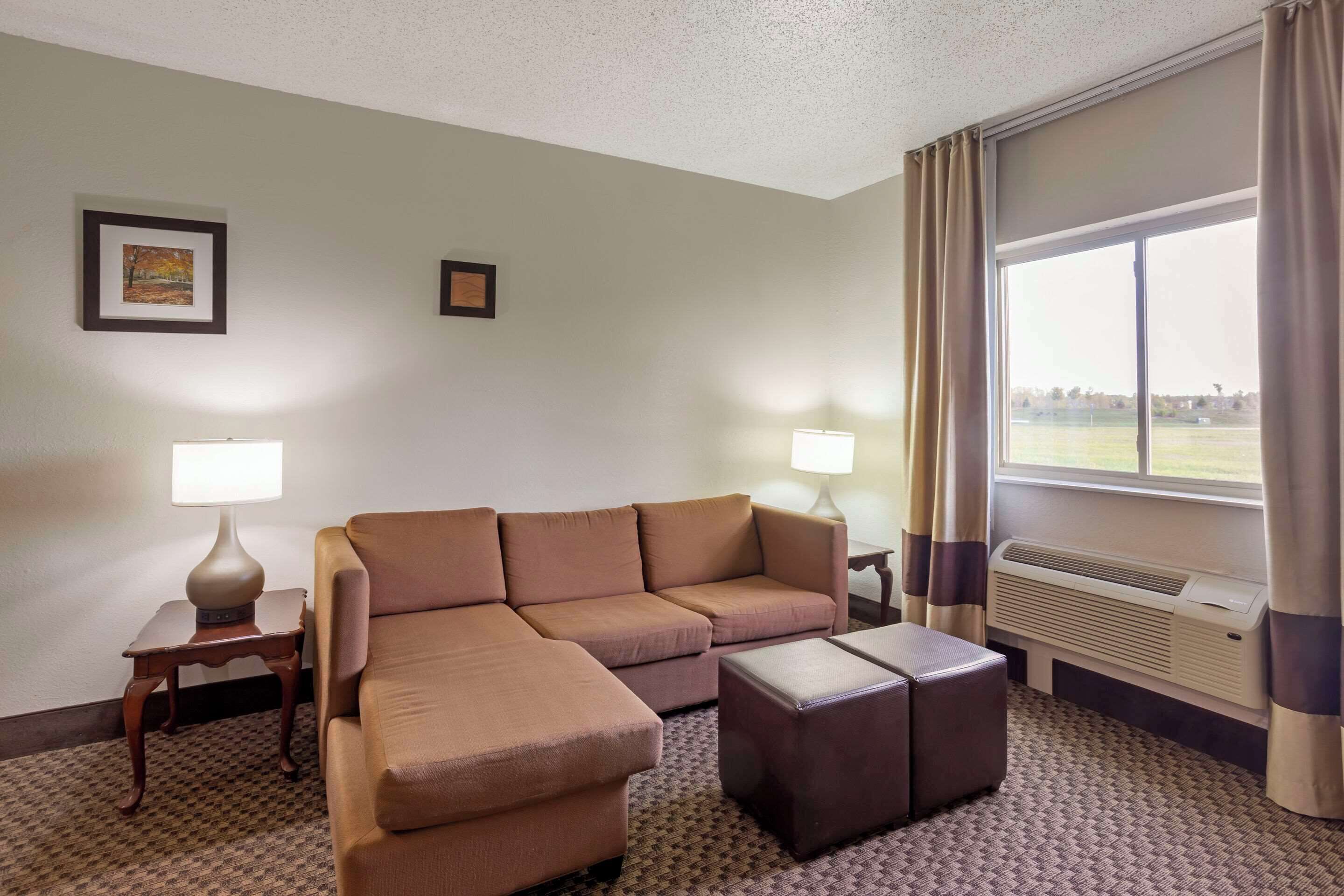 Habitación de hotel con sofá modular, una mesita de noche con lámpara, ventana con cortinas y aire acondicionado instalado en la pared.