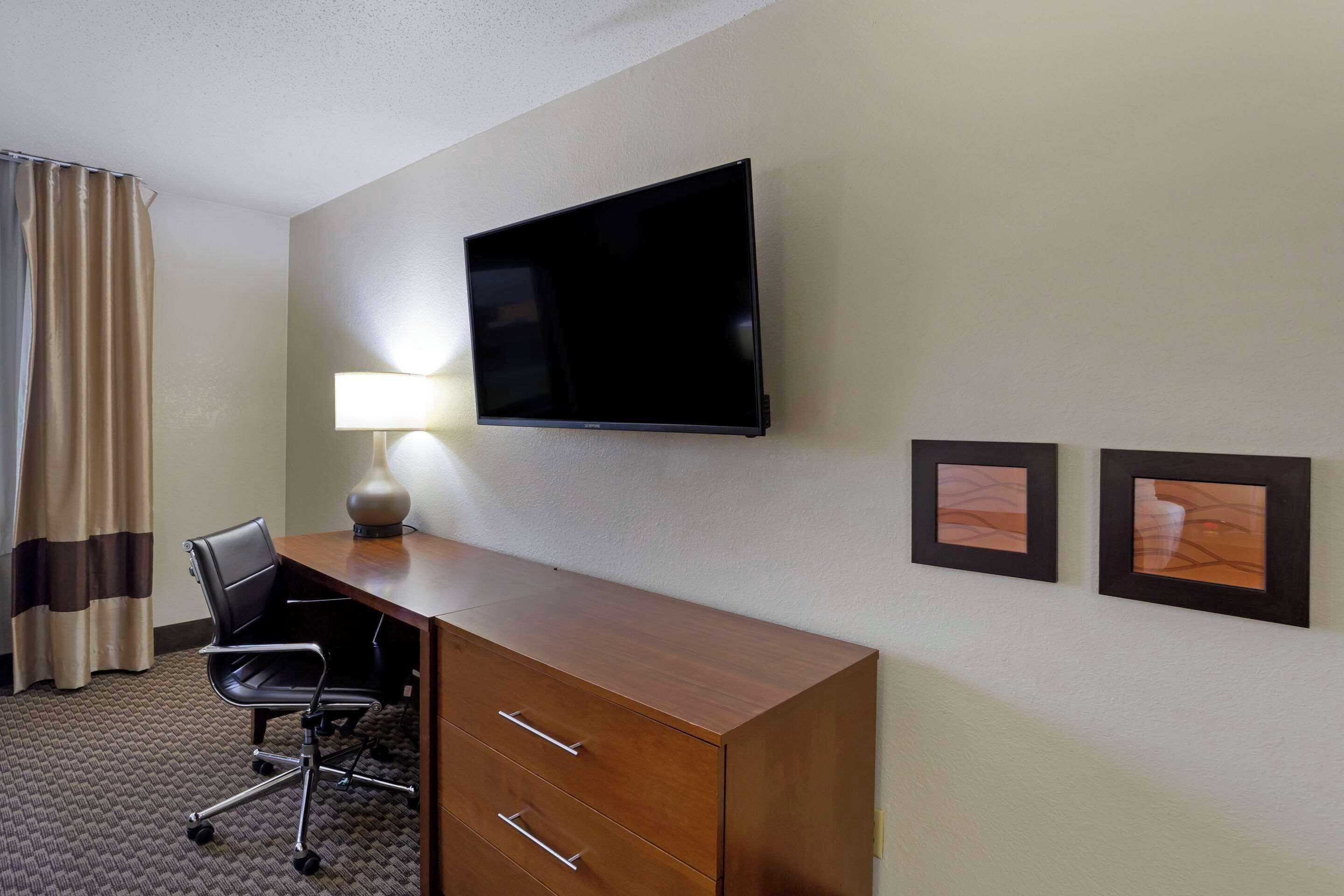 A hotel room with a flat-screen TV mounted on the wall, a wooden desk with drawers, a black office chair, a lamp, and two framed pictures on the wall.