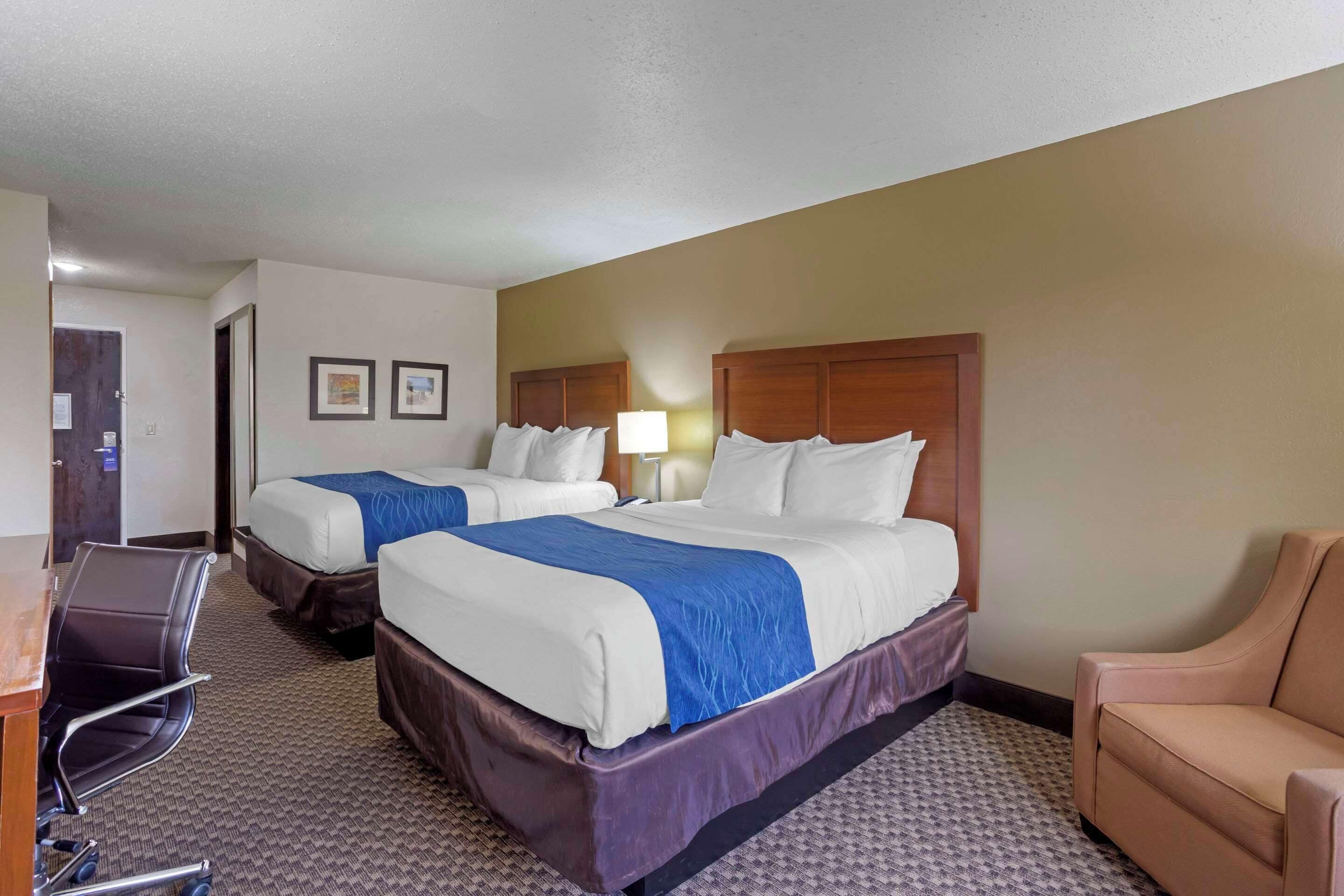 A hotel room with two beds, a desk, a chair, and a lamp.