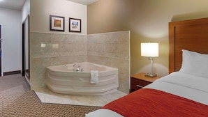 Habitación de hotel con una bañera grande, una cama con una colcha roja, una mesita de noche con una lámpara y dos cuadros en la pared.