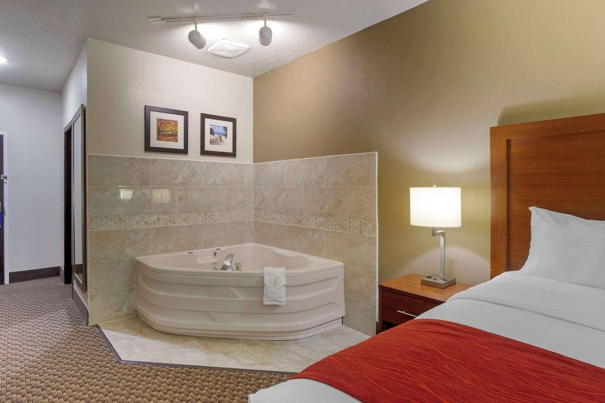 A hotel room with a large bathtub, a bed with a red blanket, a nightstand with a lamp, and two framed pictures on the wall.