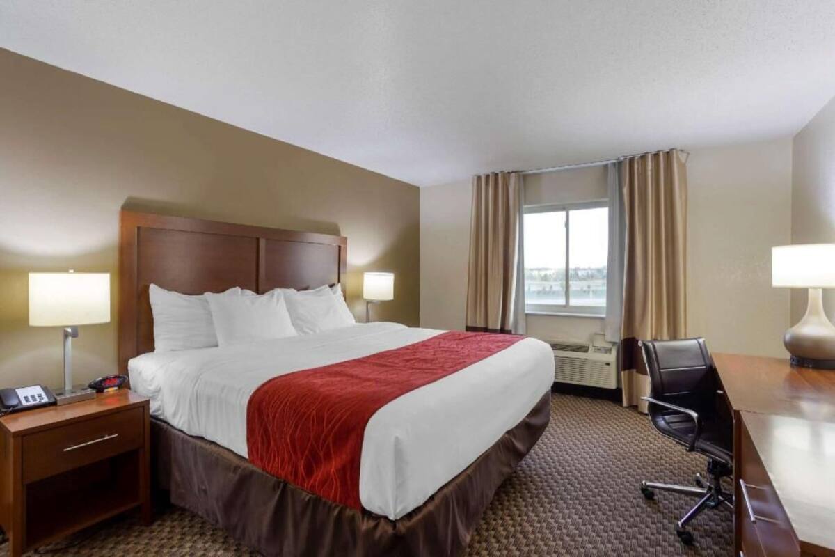 A hotel room with a large bed, a desk, a chair, a nightstand, a lamp, and a window with curtains.
