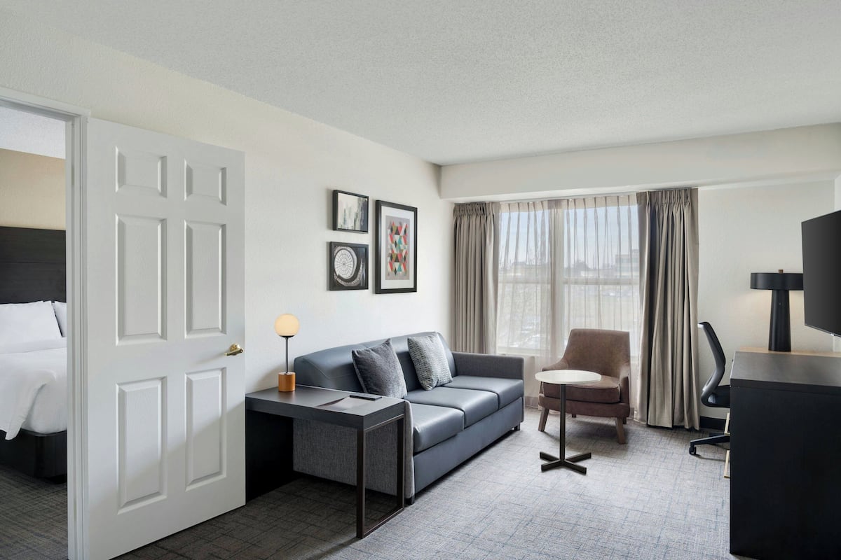 Hotelkamer met een bed, een grijze bank, een kleine tafel, een stoel, een televisie en een raam met gordijnen.