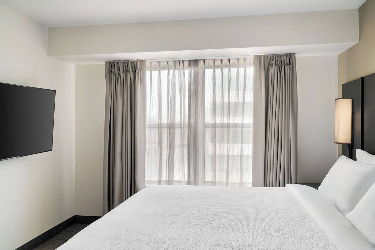 Een hotelkamer met een groot bed, een flatscreen televisie aan de muur, een bedlampje en een raam met dunne gordijnen.