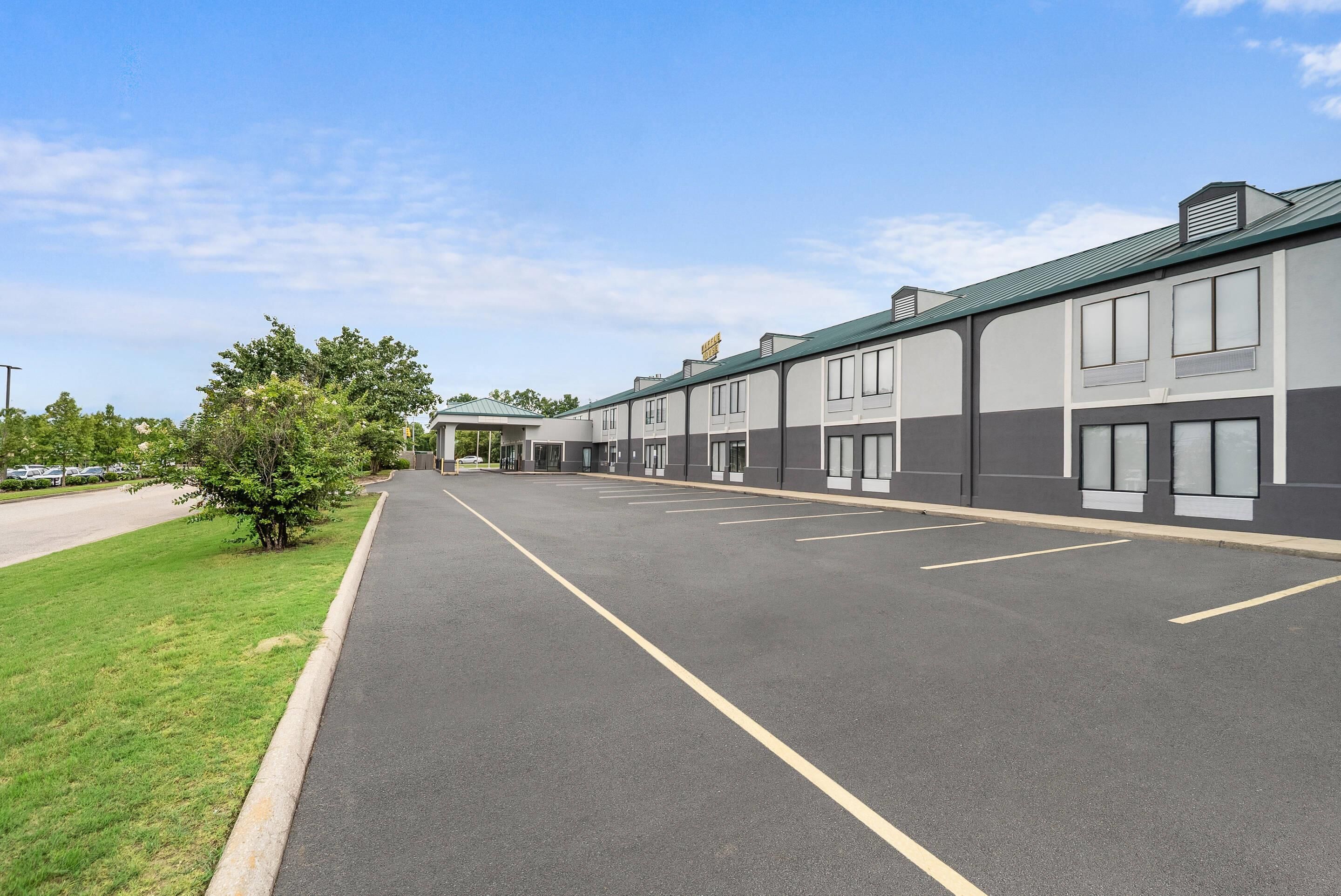 A parking lot with a row of buildings, a grassy area, and a clear sky.