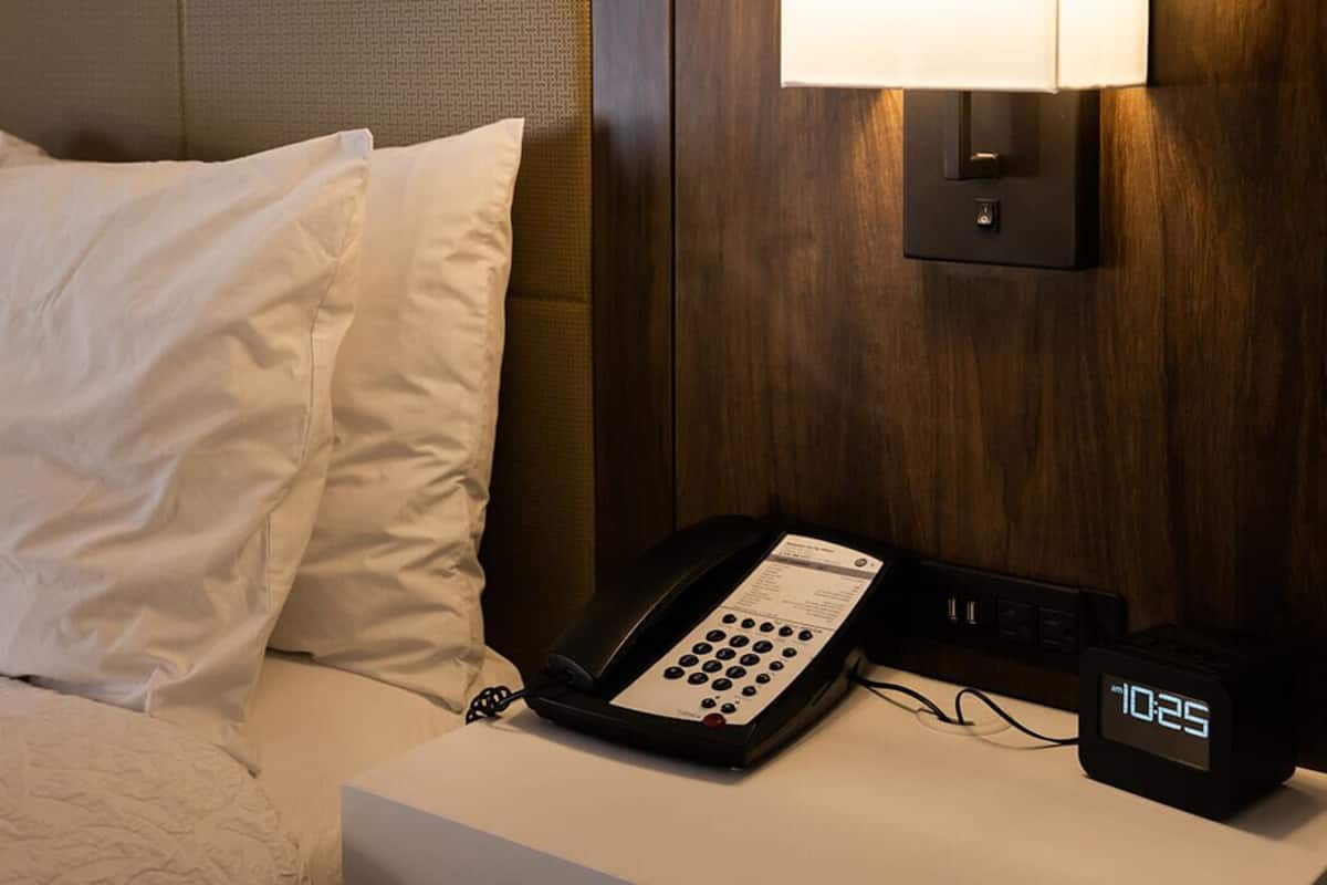 A hotel room with a bed, a bedside table with a telephone and a digital clock, and a wall-mounted lamp.