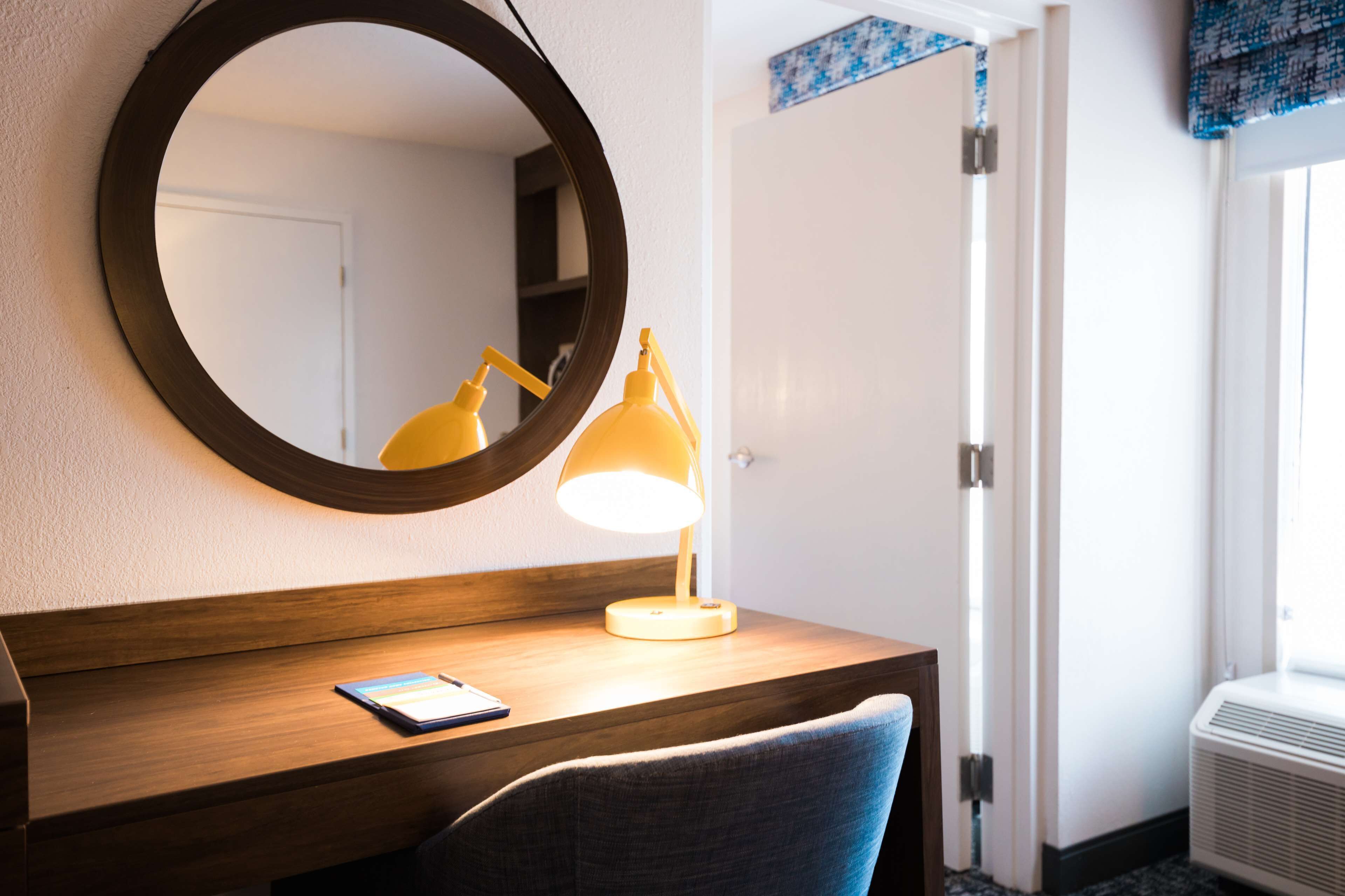 A hotel room with a wooden desk, a round mirror, a yellow desk lamp, and a chair.