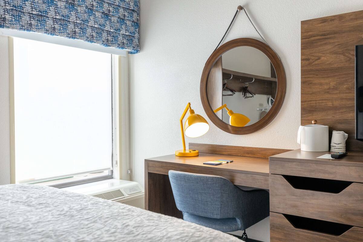 A hotel room with a wooden desk, a blue chair, a round mirror, and a bedside table with a lamp and a remote control.