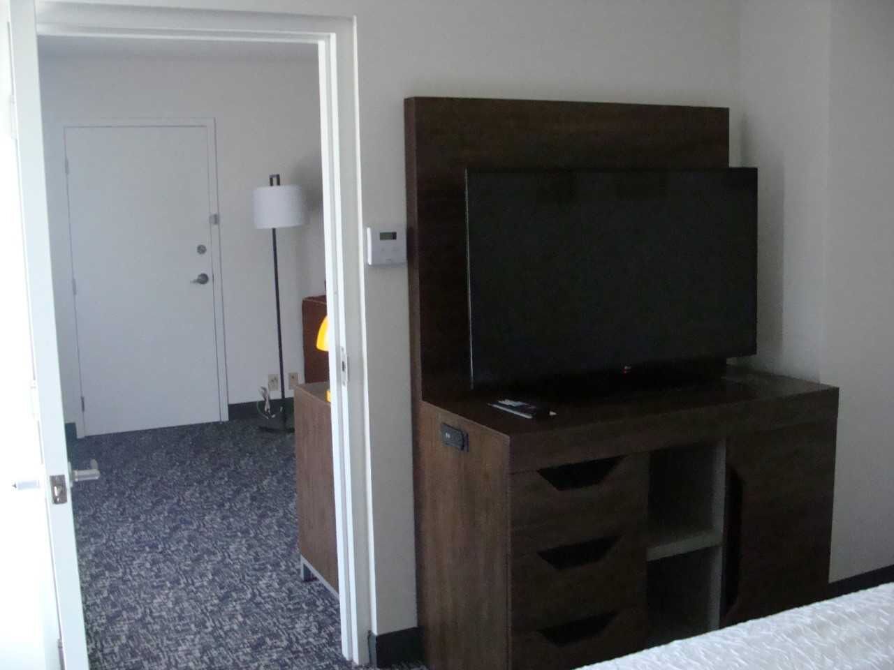 A hotel room with a dark wooden TV stand, a flat-screen TV, a lamp, and a door leading to another room.