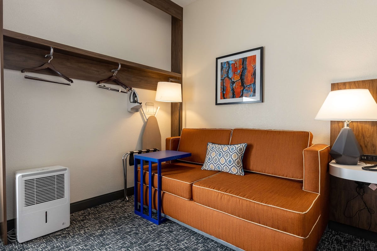 A hotel room with a brown sofa, a small table, a heater, a lamp, and a framed abstract painting on the wall.