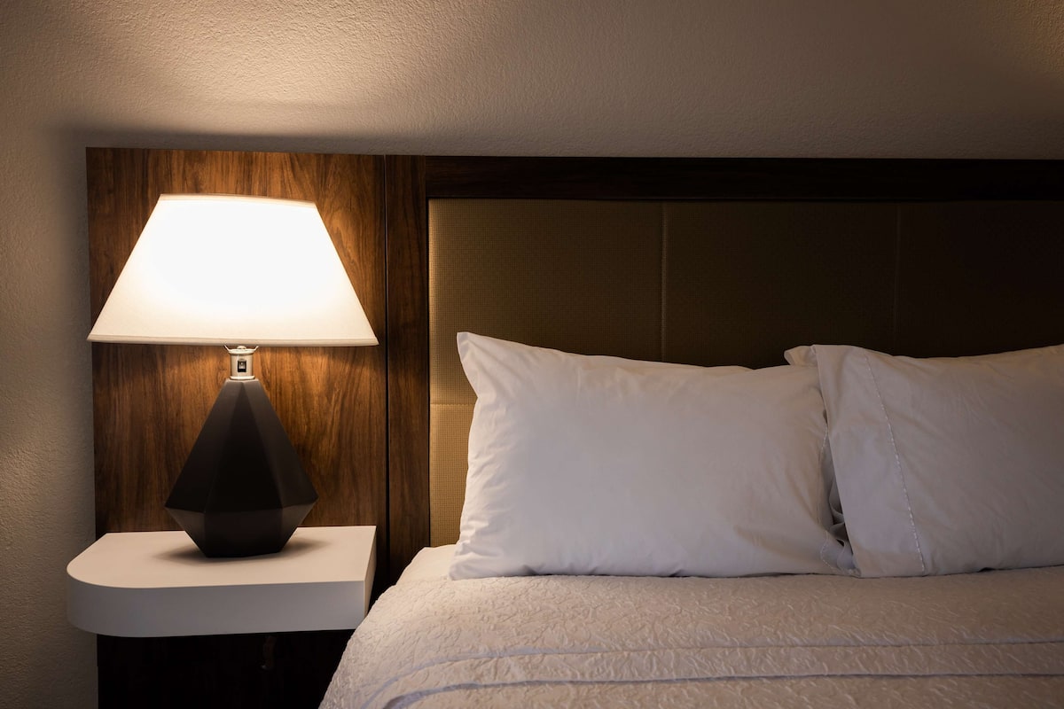A neatly made bed with white linens and pillows, a modern geometric lamp on a bedside table, and a wooden headboard.