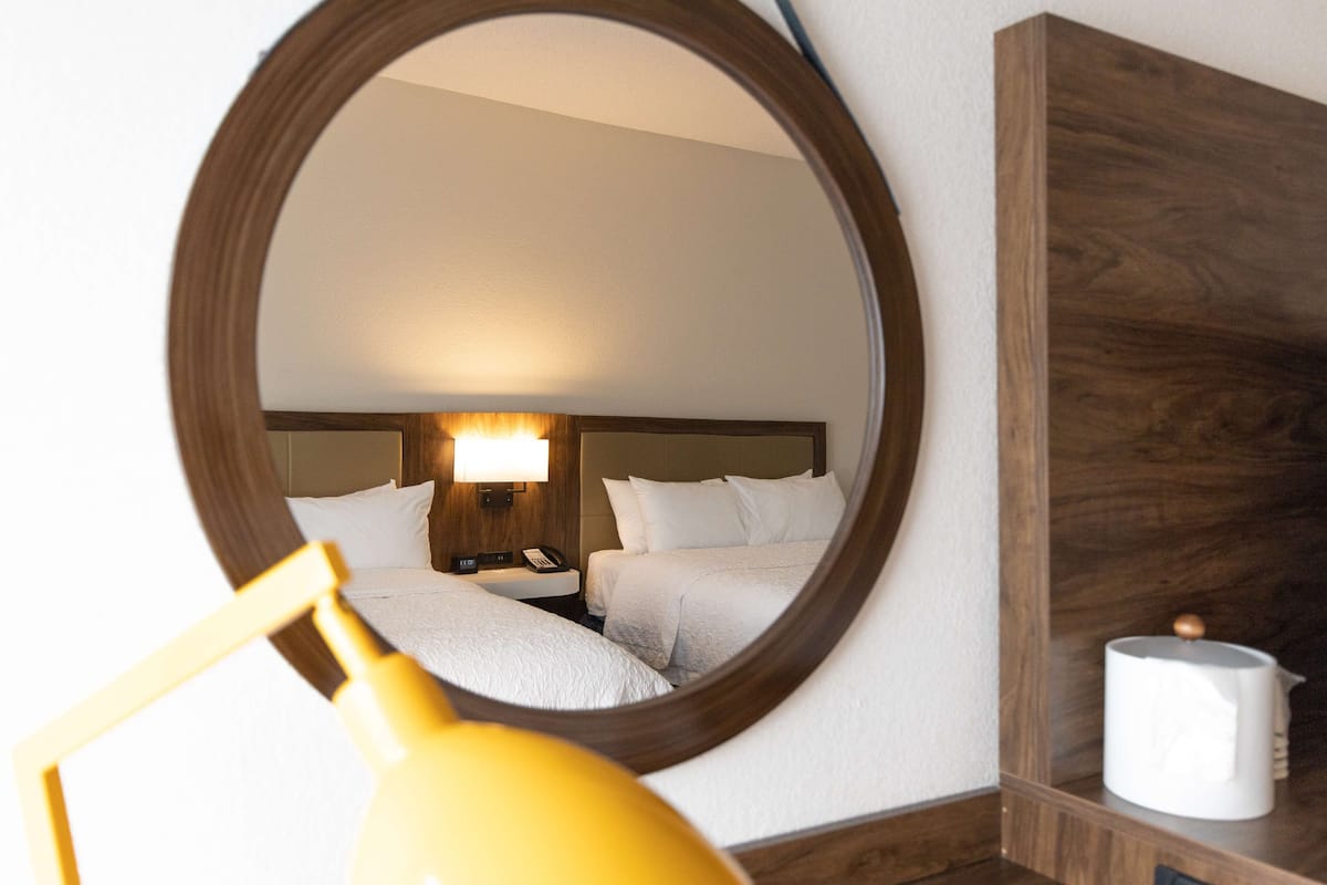 A hotel room with a round mirror reflecting a bed and a nightstand with a lamp.