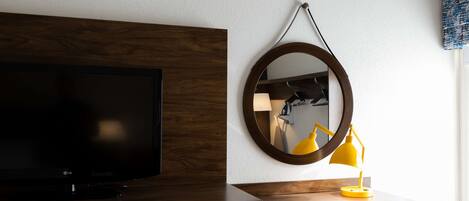 Une chambre d’hôtel avec un bureau en bois, une télévision, un miroir et une lampe de bureau jaune.