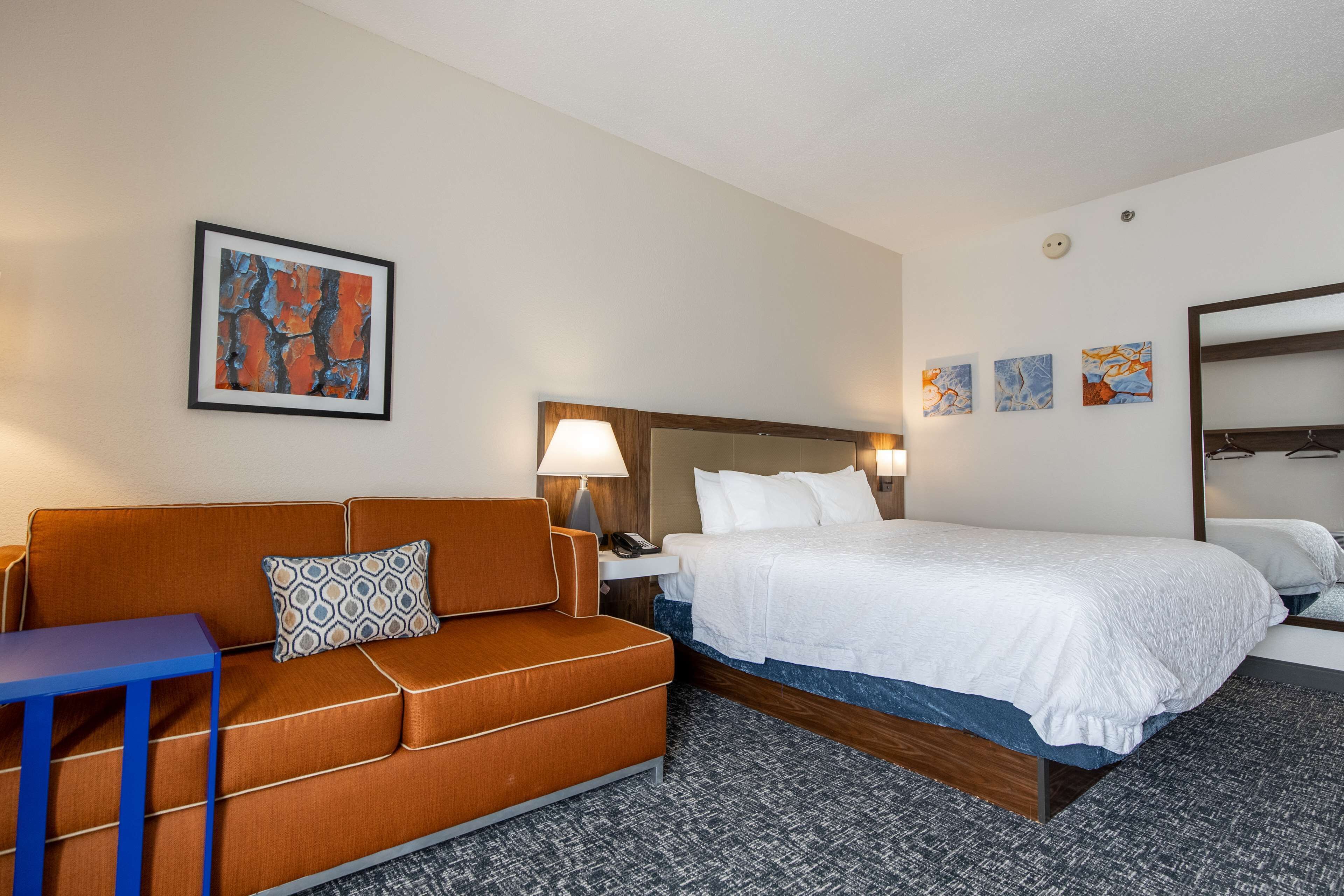 Habitación de hotel con cama, sofá, mesita de noche, lámpara y obras de arte en las paredes.