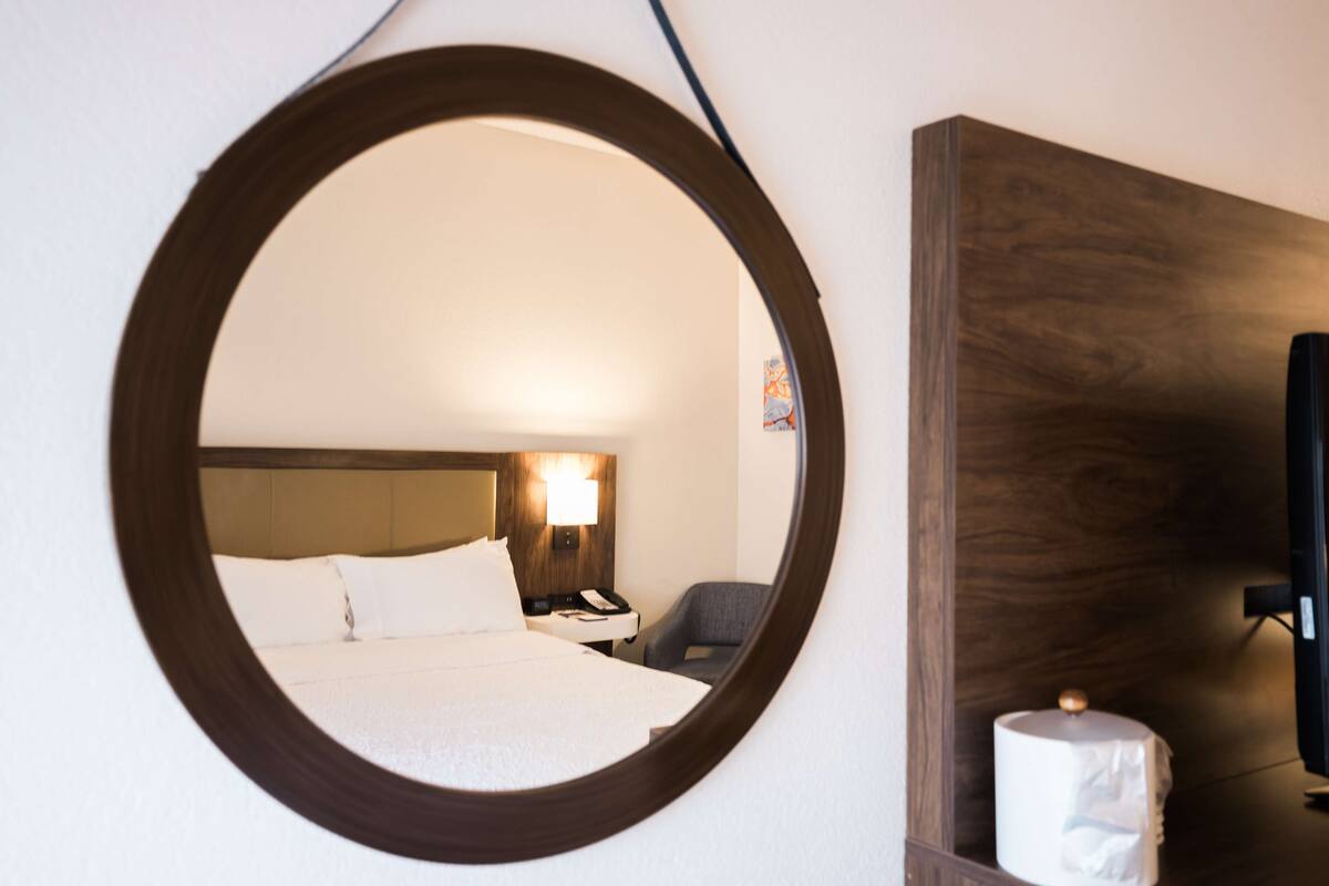A hotel room with a large round mirror, a bed with white linens, a wooden headboard, a bedside table with a lamp, a chair, and a television mounted on the wall.