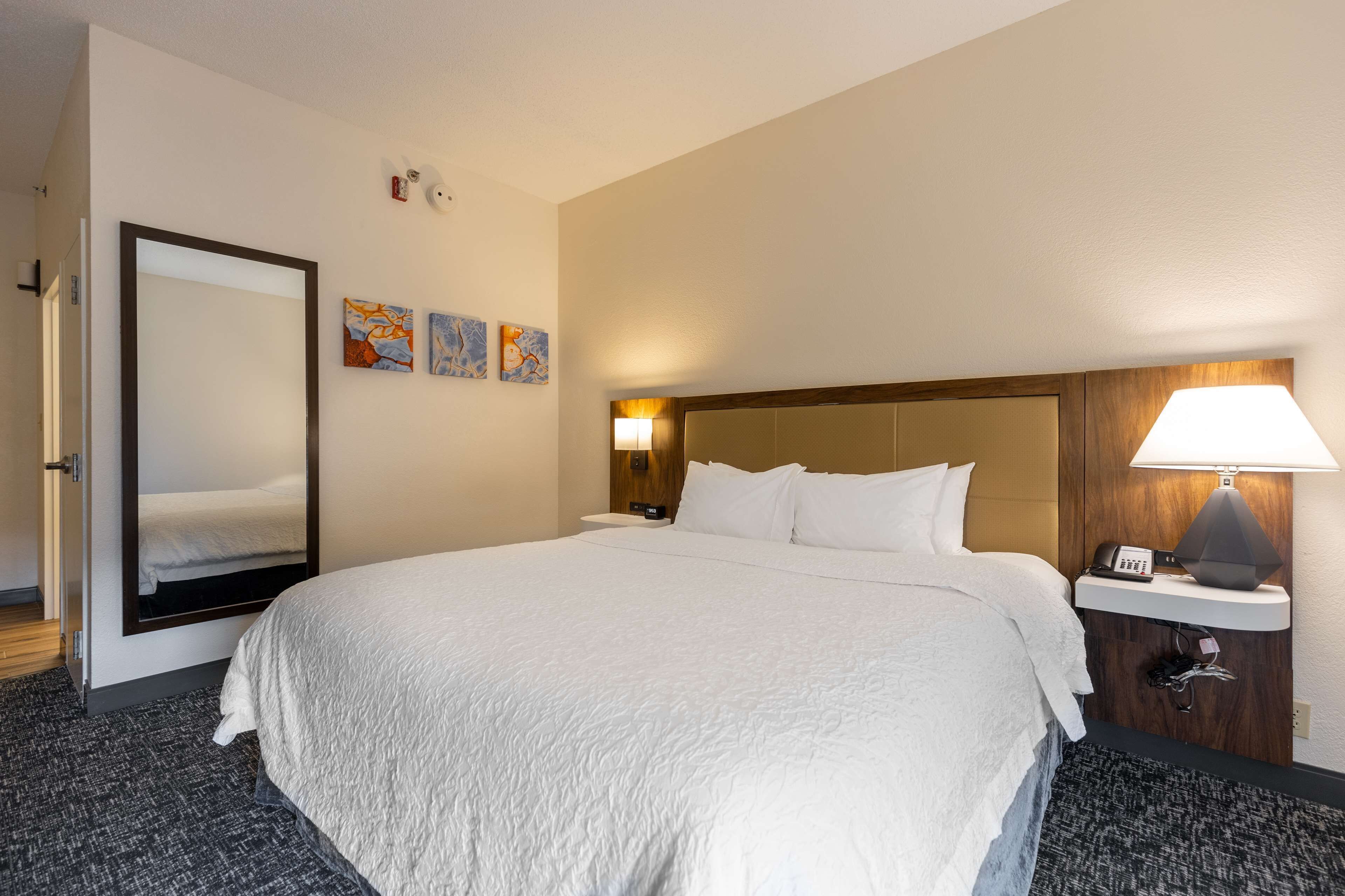 A hotel room with a large bed, a mirror, a nightstand with a lamp, and three artwork pieces on the wall.