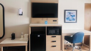 Hotellihuone, jossa on taulutelevisio, työpöytä tuolilla, jääkaappi, mikroaaltouuni, kahvinkeitin ja seinällä kehystetty taulu.