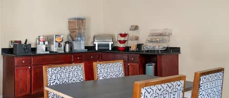 Una cocina compacta equipada con cafetera, tostadora y licuadora. Se observan muebles de madera y una mesa de comedor con sillas.