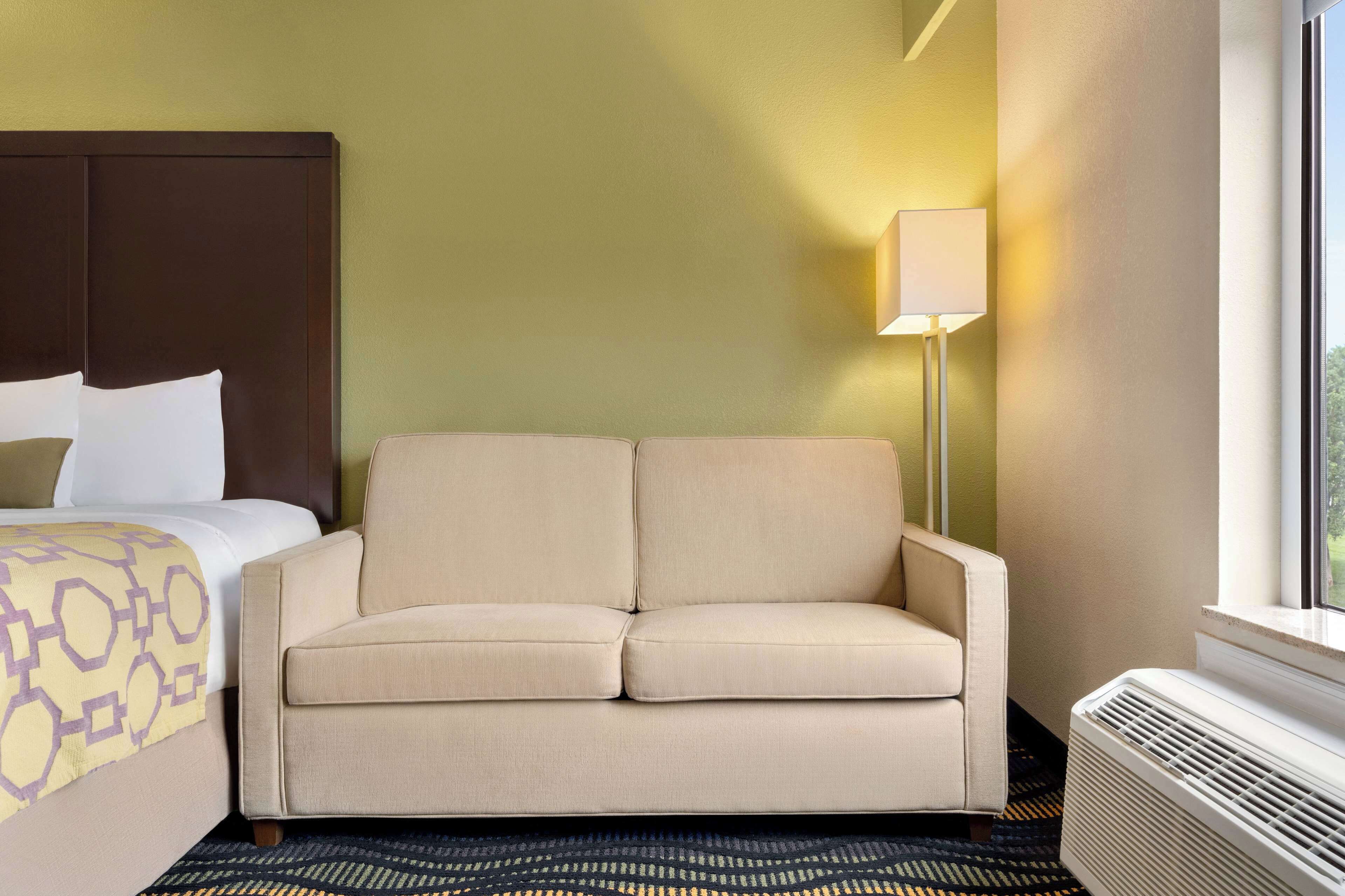 A hotel room with a bed, a beige sofa, a standing lamp, and a window.