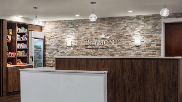 A reception area with a stone wall, a Baymont Inn & Suites sign, a refrigerator stocked with beverages, and a wooden counter. - Baymont by Wyndham Georgetown/Lexington North (Georgetown)