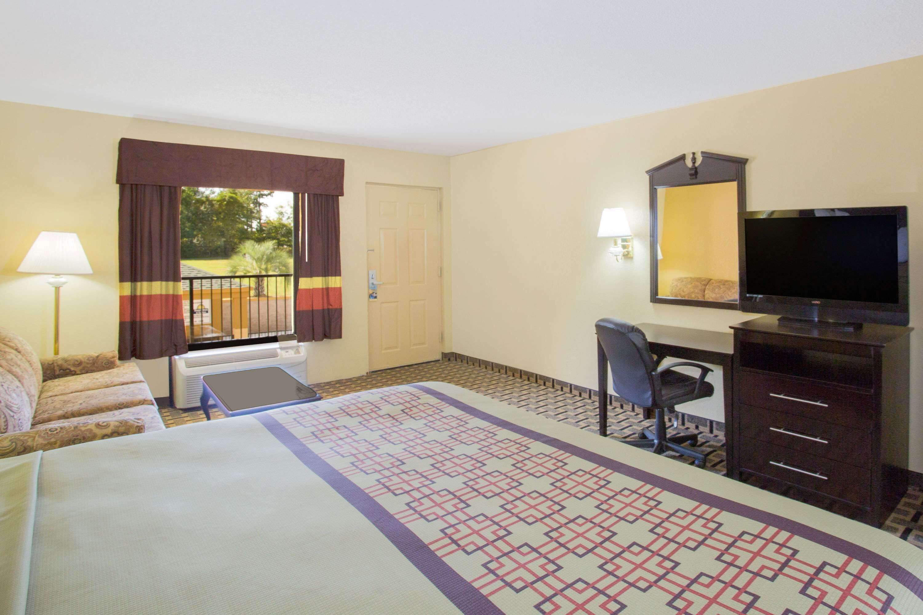 Una habitación de hotel con cama, escritorio, silla, televisor y ventana con cortinas.