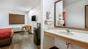Habitación de hotel con cama, escritorio, silla, televisión, espejo, lavamanos y toallas.