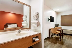 Une salle de bain d’hôtel avec un grand miroir, un lavabo, des toilettes et un espace douche. On y trouve un porte-serviettes avec des serviettes, une chaise en bois et une petite table avec un téléviseur.